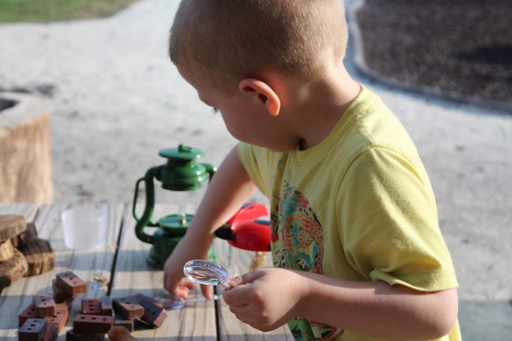 Young boy in yellow shirt learns STEM skills, playing outside with magnifying glass, building blocks and a green lantern.